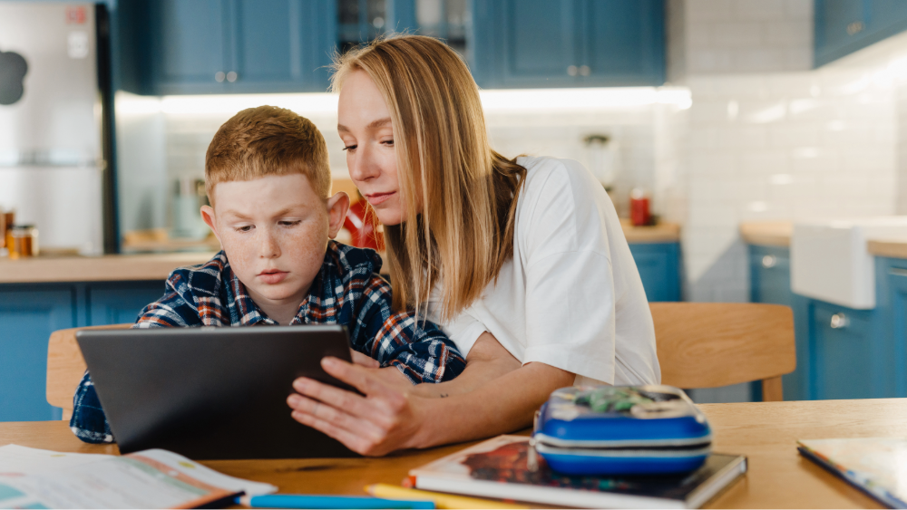 A parent and a primary school child sit in the kitchen using a tablet surround by school supplies