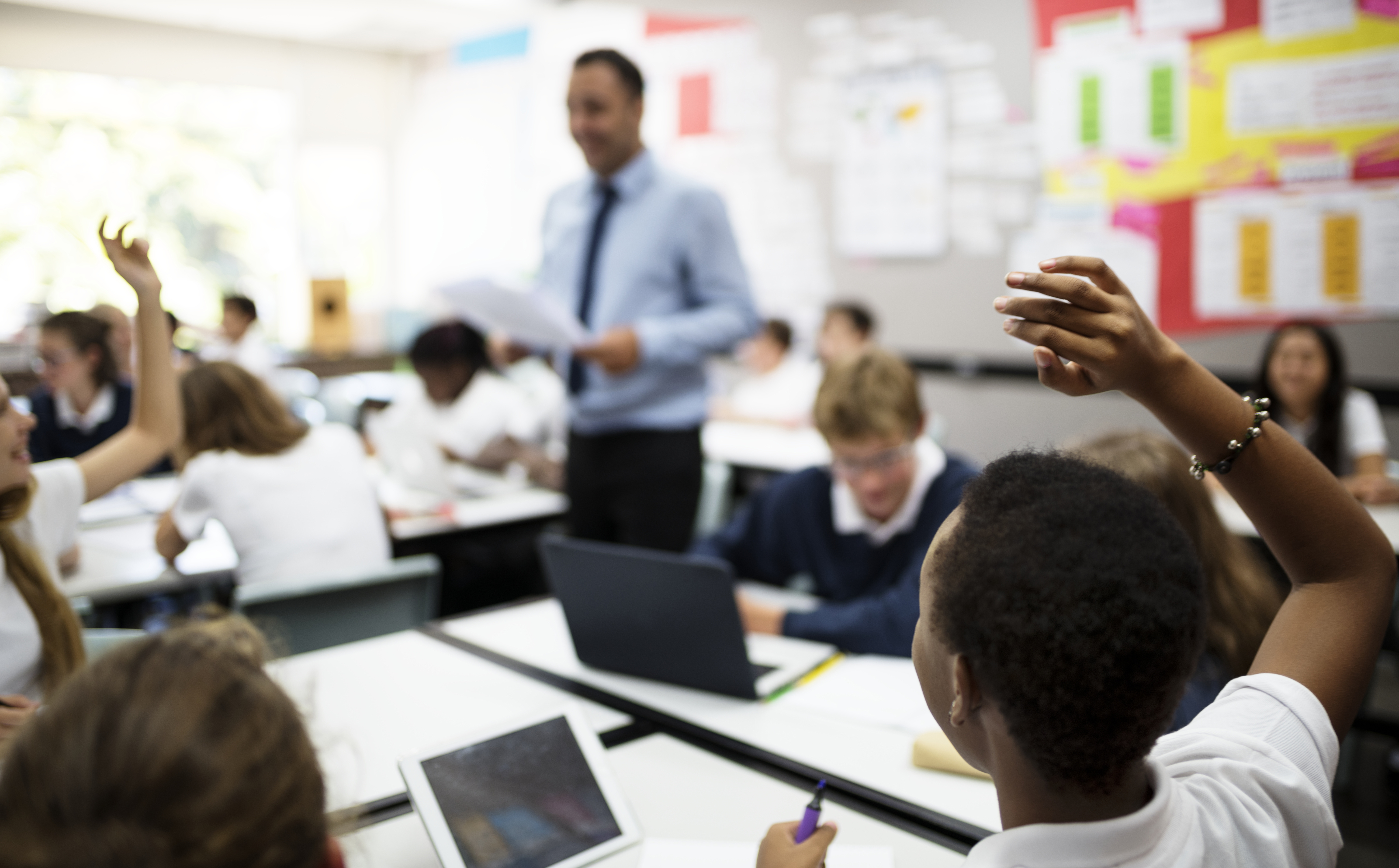 A classroom full of secondary school students working on tablets with the teacher walking between the desks in the background