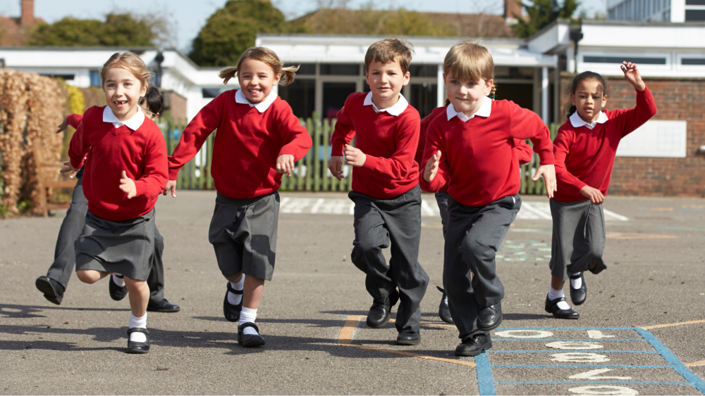 Seven primary school students in uniforms with red jumpers run happily through the playground towards the camera