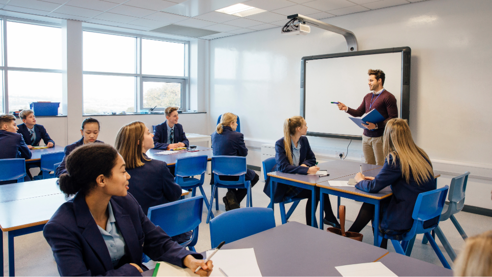 A teacher stands in front of a smart board and projector in a secondary school classroom filled with students.