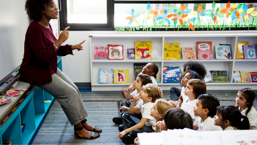 Young students sit on the carpet in front of their primary school teacher as she talks, there are books on the shelf behind them and colourful flowers made by the students stuck on the windows