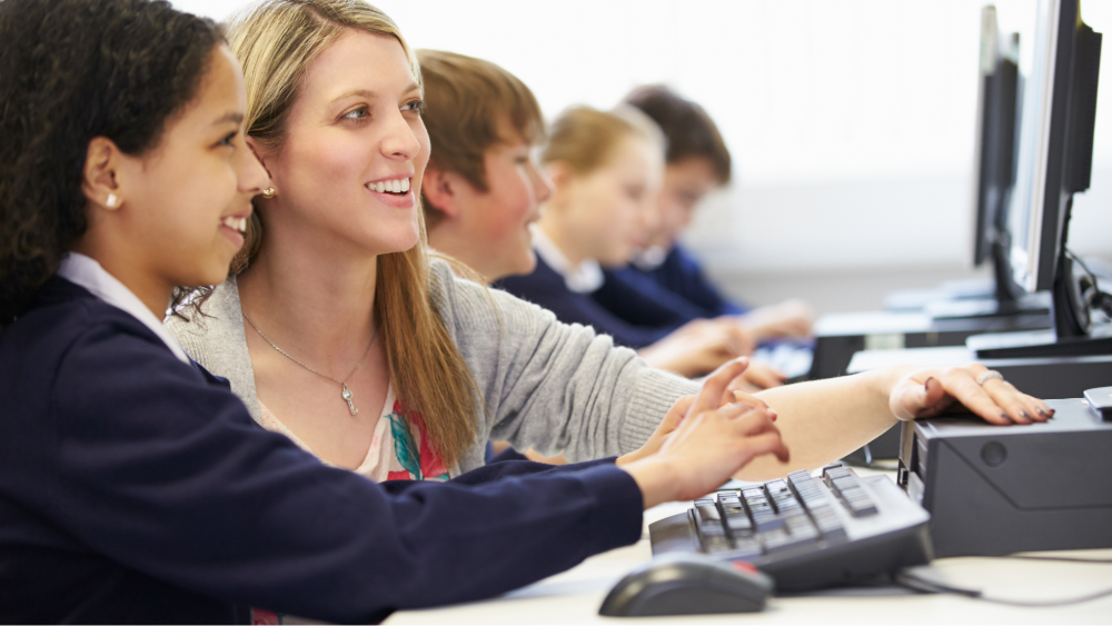 A teacher and secondary student work together on a desktop computer in ICT class