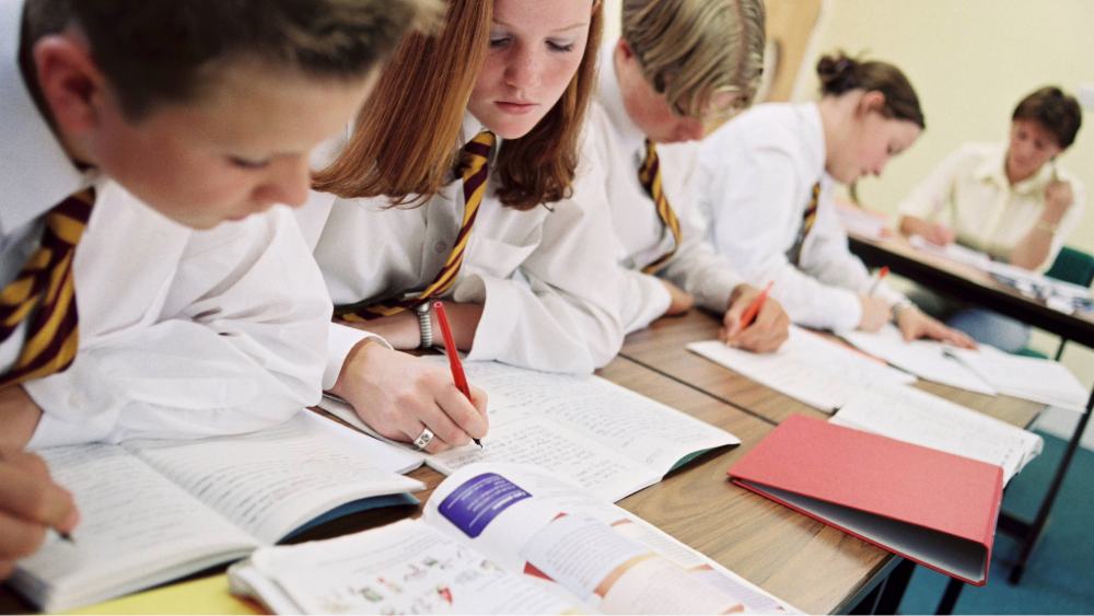 Four secondary students sitting at desks in a classroom make notes from a textbook