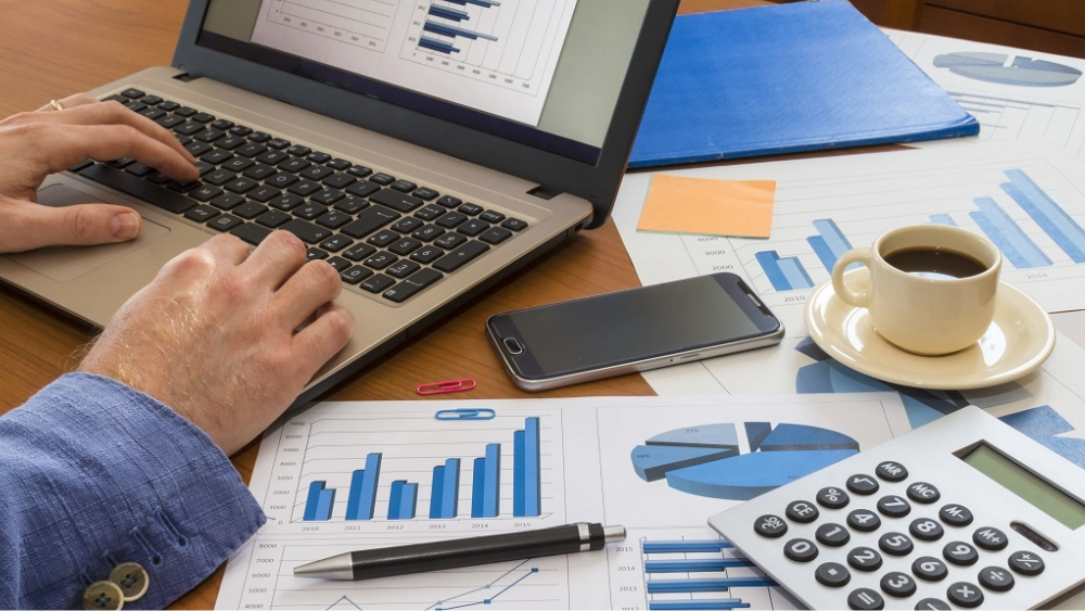 A finance manager works on his laptop while the desk is covered in insight reports, a phone, calculator and a cup of coffee.