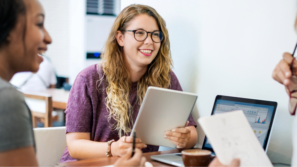 Teachers sit around a laptop having a meeting, one teacher is holding a tablet, smiling and laughing