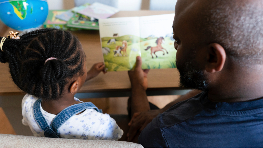 A young child and her parent read a book together at the dining table