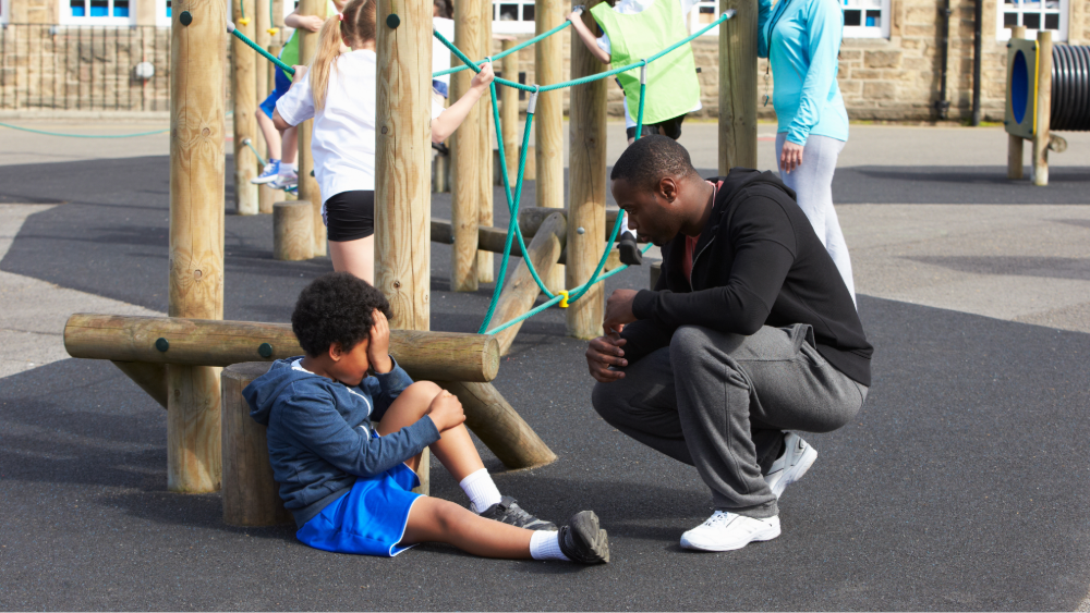 A child is holding his knee on the playground while a teacher checks on him