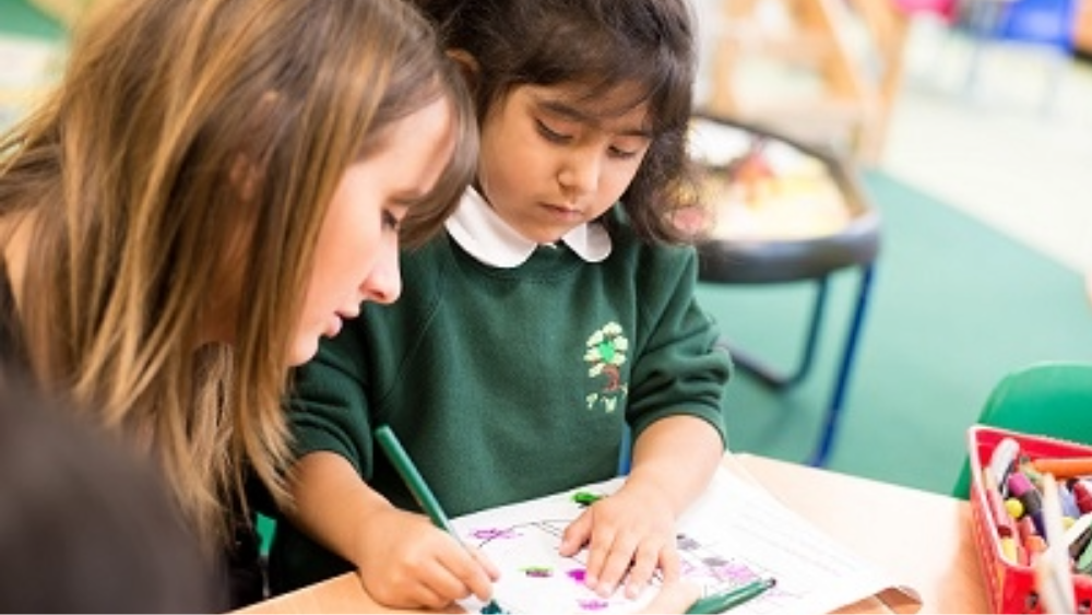 Primary school student colours in a drawing while sitting next to her teacher